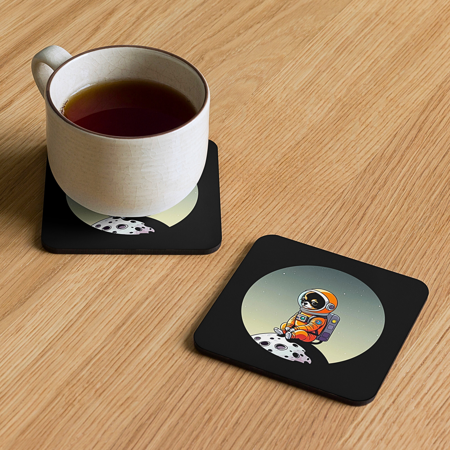 Photo of a square black coaster printed with a cute illustration of a Chihuahua dog wearing an astronaut suit sitting on a small moon. Next to it there is another coaster with a cup of tea on it. Both are laying on top of a wooden surface.